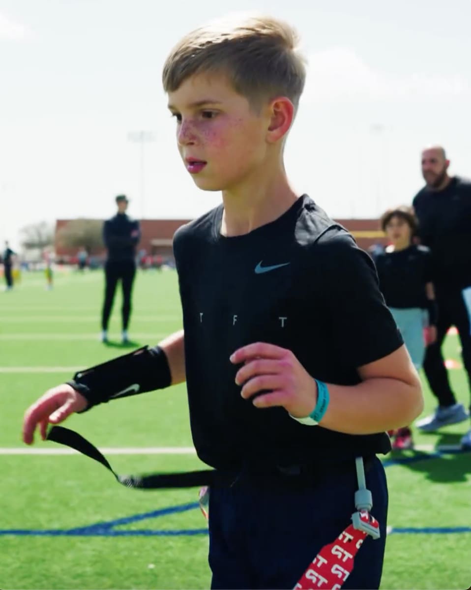 A little boy, Theo, who wore a black Nike shirt and a naval pants, concentrated intensively on the flag football field. He has a determined expression with a bracelet and a flag grinder that is ready.