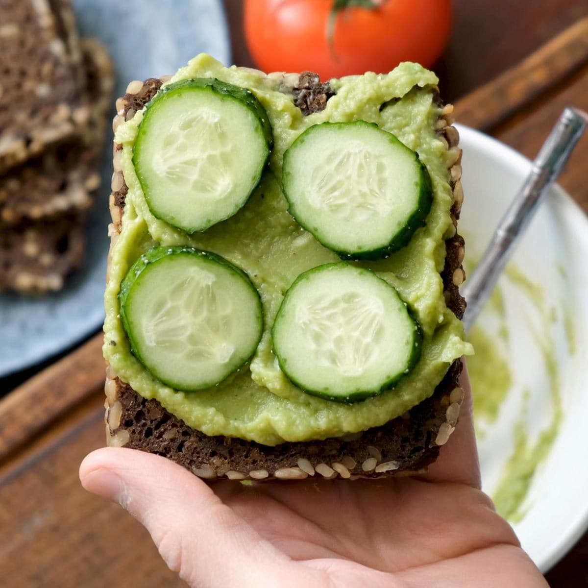 Male hand holding vegan rye bread toast with mashed avocado and cucumber slices
