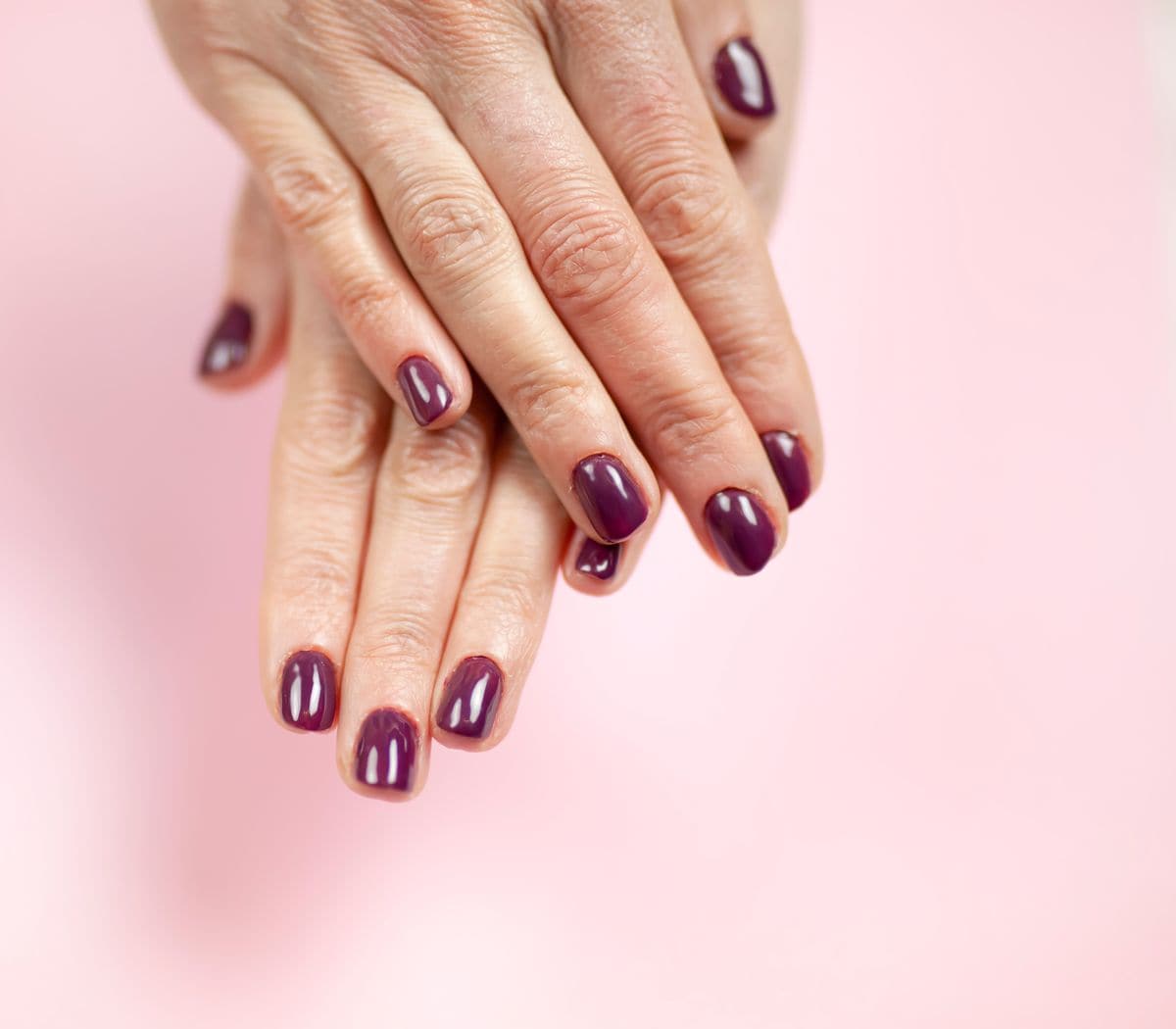 Close-up of hands with short, glossy plum-colored nails against a soft pink background