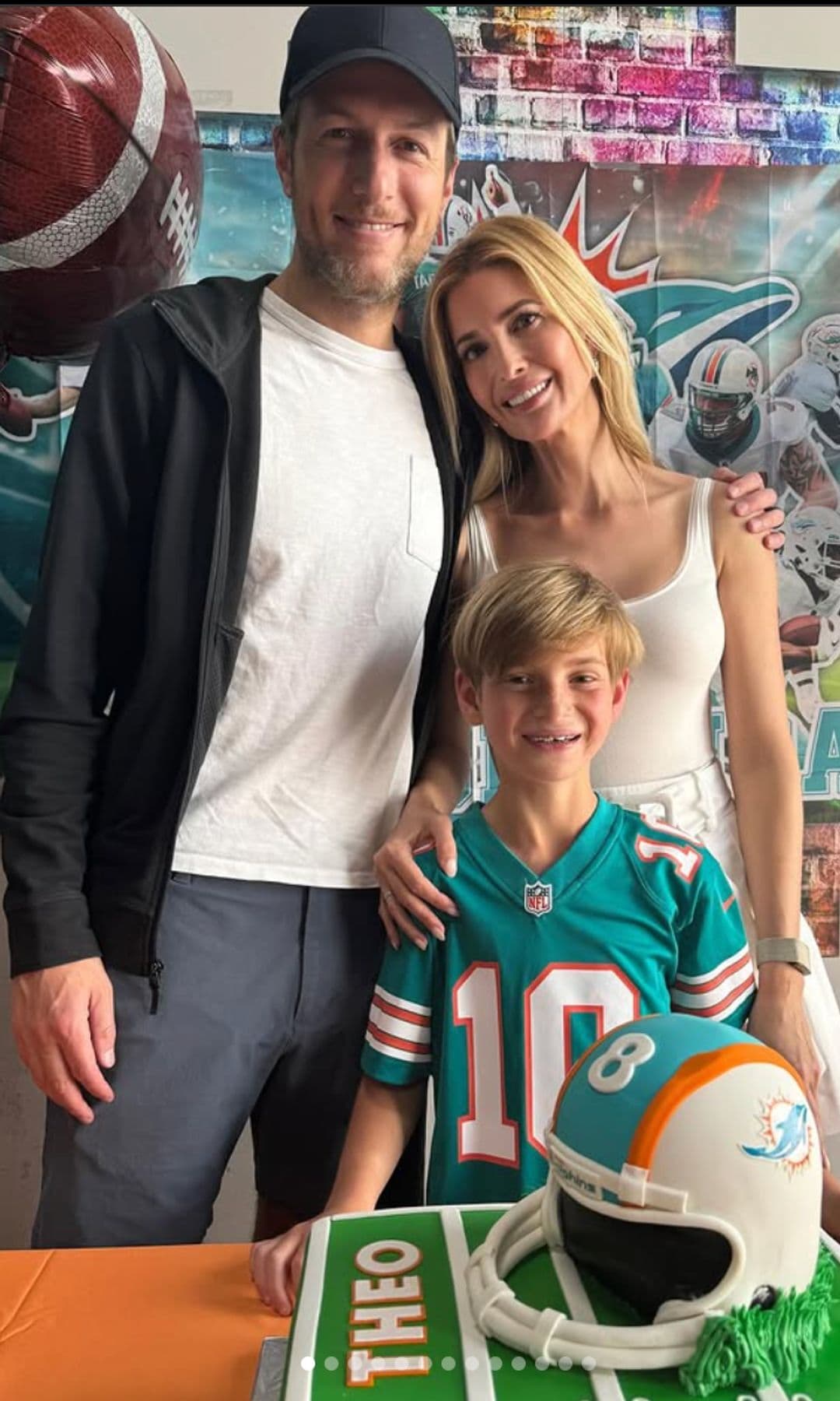 Jared, Ivanka, and Theo pose together at Theo’s Miami Dolphins-themed birthday party. Theo wears a Dolphins jersey and stands in front of a custom football-themed cake featuring his name and a helmet decoration.