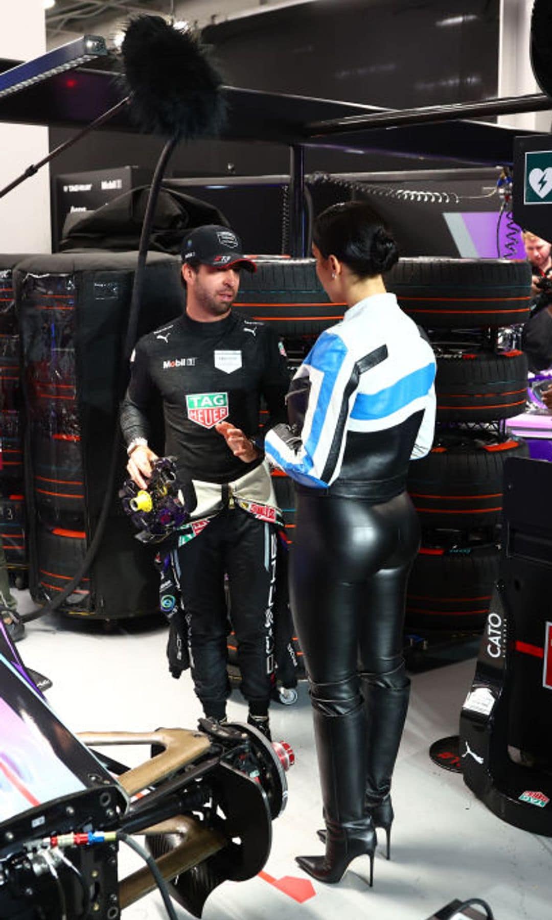 Georgina Rodríguez in a black leather outfit with a blue and white racing-style jacket, talking to a Formula E driver in the pit area.